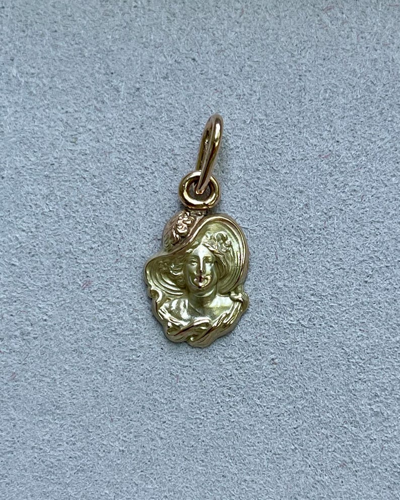 May include: A gold-colored pendant featuring a woman's face wearing a wide-brimmed hat. The pendant has intricate details, including floral accents on the hat. The bail is a simple loop for attaching to a chain. The pendant is likely made of gold.
