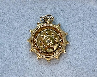 May include: A gold pendant with a detailed, circular design. The pendant features a central starburst pattern with a small, clear gemstone. The outer edge has a series of small, rounded protrusions. The pendant is suspended from a small loop.