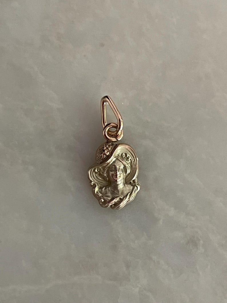 May include: A silver-toned pendant featuring a woman's profile wearing a wide-brimmed hat. The pendant has a detailed, Art Nouveau style design with a loop for attaching to a chain. The woman's face is visible, and the hat has decorative elements.