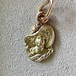 May include: A gold-colored pendant featuring a woman's profile wearing a wide-brimmed hat. The pendant has a detailed design with floral accents and a loop for a chain. The pendant is approximately 0.75 inches in height.