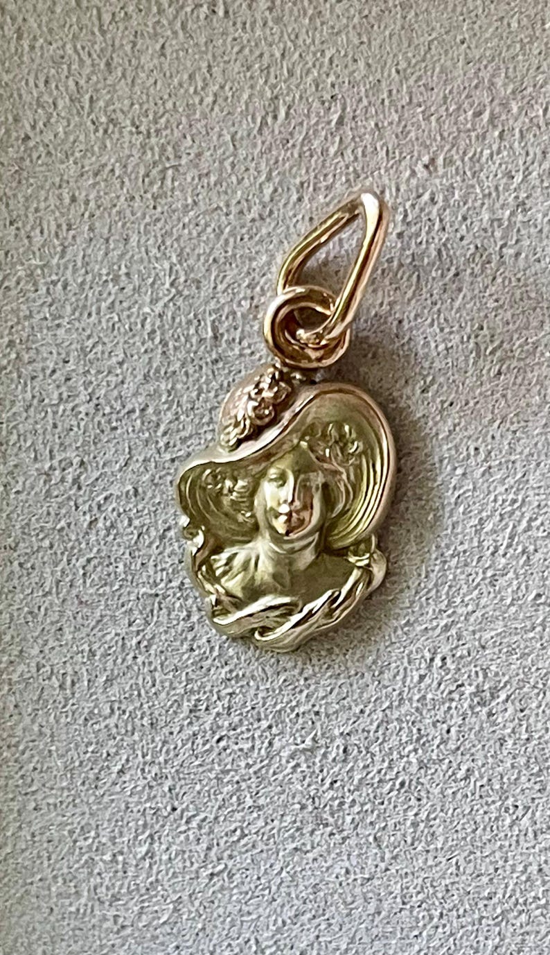 May include: A gold-colored pendant featuring a woman's profile wearing a wide-brimmed hat. The pendant has a detailed design with floral accents and a loop for a chain. The pendant is approximately 0.75 inches in height.