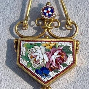 Puede incluir: Colgante de oro con dise&ntilde;o floral de mosaico. El colgante presenta un mosaico en forma de escudo con flores blancas, rosas, azules y amarillas. El mosaico est&aacute; enmarcado en oro y tiene un peque&ntilde;o mosaico redondo en la parte superior.