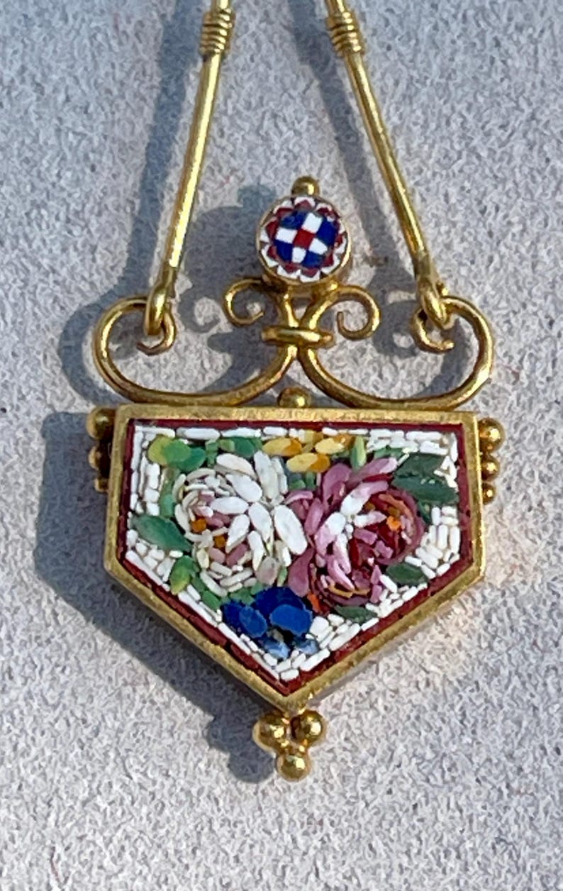 Puede incluir: Colgante de oro con dise&ntilde;o floral de mosaico. El colgante presenta un mosaico en forma de escudo con flores blancas, rosas, azules y amarillas. El mosaico est&aacute; enmarcado en oro y tiene un peque&ntilde;o mosaico redondo en la parte superior.