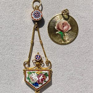 Puede incluir: Dos colgantes de oro. Uno es un dise&ntilde;o largo y ornamentado con un mosaico floral. El otro es un colgante redondo con un dise&ntilde;o de flor rosa. Ambos tienen lazos dorados para colgar.