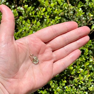 May include: A small, gold-colored pendant in the shape of a flower or seashell. The pendant is held in an open palm against a backdrop of green foliage. The pendant has intricate details and a small loop for a chain.