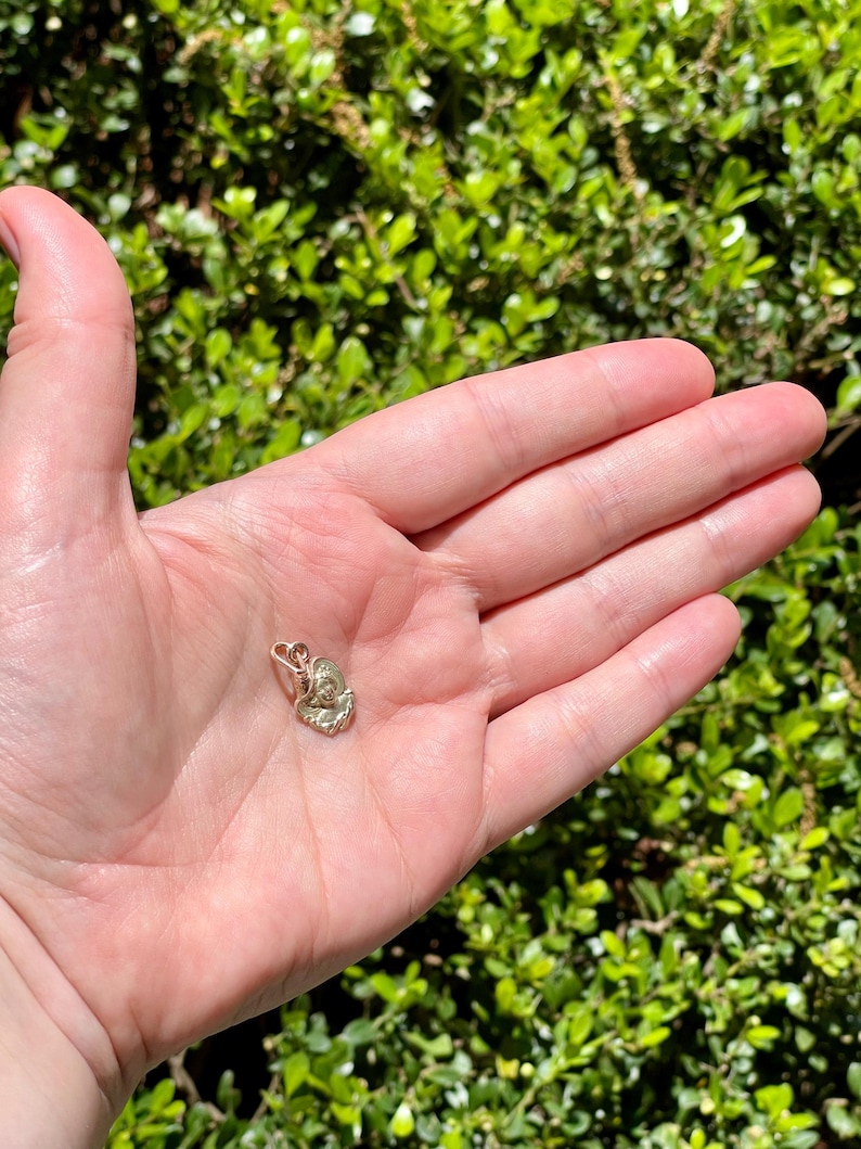 May include: A small, gold-colored pendant in the shape of a flower or seashell. The pendant is held in an open palm against a backdrop of green foliage. The pendant has intricate details and a small loop for a chain.