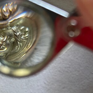 May include: Close-up of a silver-toned metal object with a detailed relief of a classical female face and floral elements. The object has a reflective surface and appears to be a decorative piece or jewelry component.