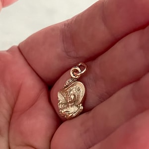 May include: A small, gold-colored pendant featuring a detailed relief of a woman's face wearing a hat. The pendant has a loop at the top for attaching to a chain or bracelet. The design is intricate and shows fine details.
