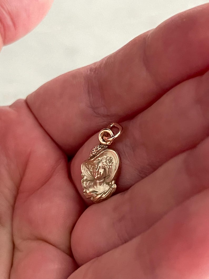 May include: A small, gold-colored pendant featuring a detailed relief of a woman's face wearing a hat. The pendant has a loop at the top for attaching to a chain or bracelet. The design is intricate and shows fine details.