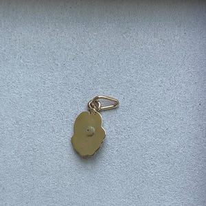 May include: A gold-colored charm with a flower-like shape and a small loop for attaching to a necklace or bracelet. The charm has a textured surface and a small, round detail in the center. Jewelry.