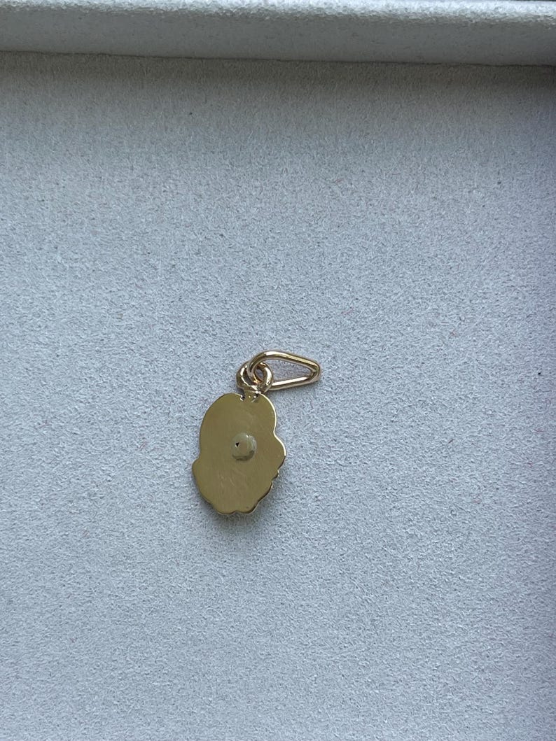 May include: A gold-colored charm with a flower-like shape and a small loop for attaching to a necklace or bracelet. The charm has a textured surface and a small, round detail in the center. Jewelry.