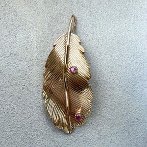 May include: A rose gold pendant shaped like a textured feather with a central spine and two small, round, vibrant pink gemstones set along the right side of the leaf. The metal surface features fine, etched lines mimicking the appearance of a feather.