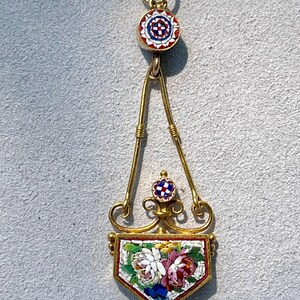 Puede incluir: Colgante dorado con dise&ntilde;o floral de mosaico. El colgante presenta un mosaico en forma de escudo con flores blancas, rosas y azules. La parte superior e inferior del colgante tienen peque&ntilde;os c&iacute;rculos de mosaico con patrones rojos, blancos y azules.