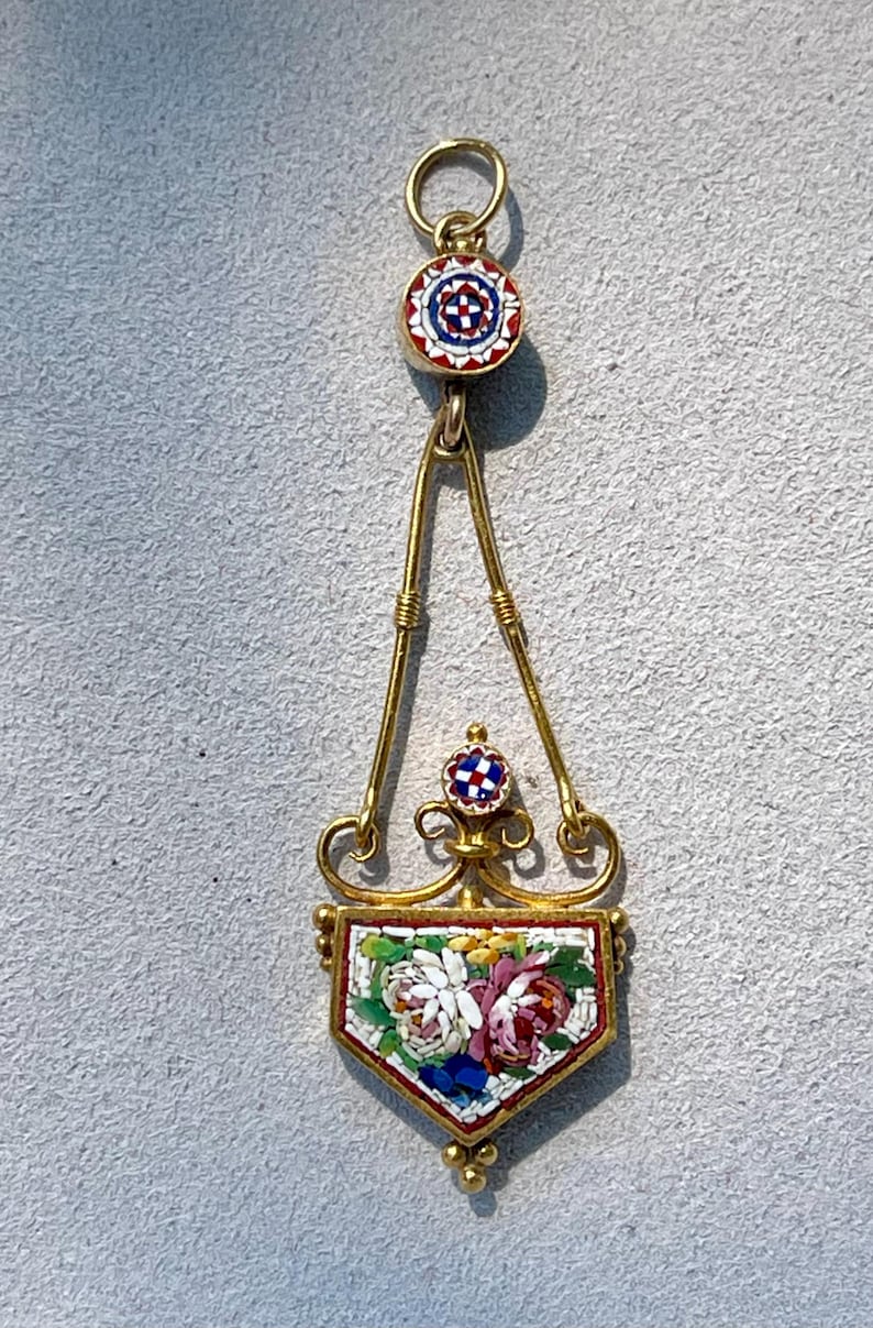 Puede incluir: Colgante dorado con dise&ntilde;o floral de mosaico. El colgante presenta un mosaico en forma de escudo con flores blancas, rosas y azules. La parte superior e inferior del colgante tienen peque&ntilde;os c&iacute;rculos de mosaico con patrones rojos, blancos y azules.