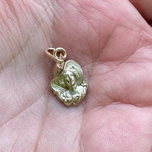 May include: A gold-colored pendant charm featuring a woman's face wearing a wide-brimmed hat. The charm has a detailed, vintage design with a loop at the top for attaching to a necklace or bracelet. The charm is small and delicate.
