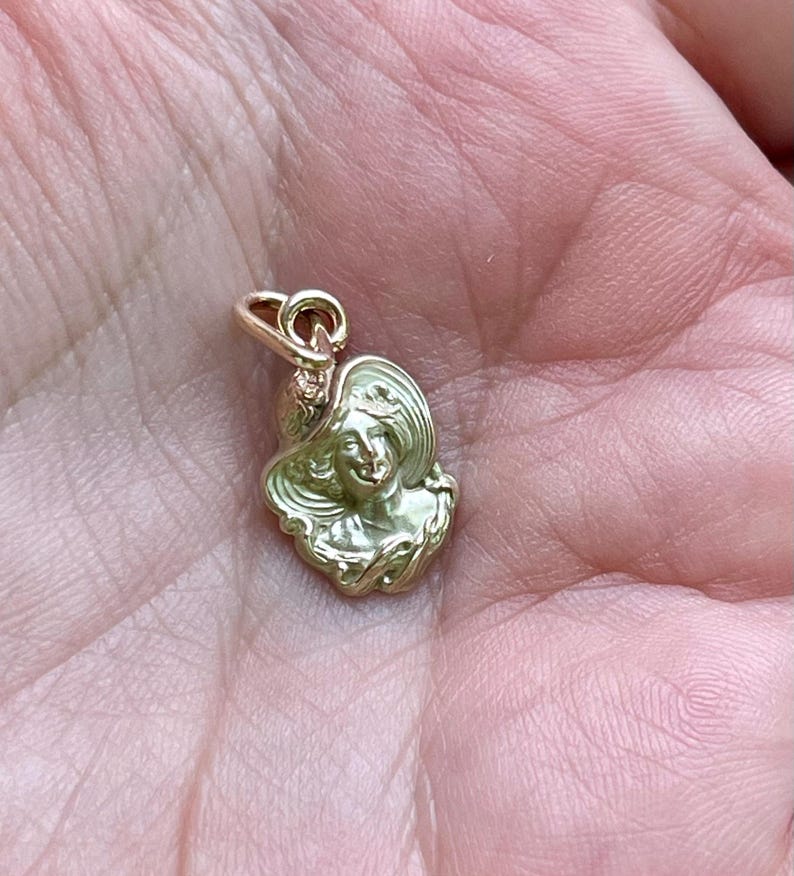 May include: A gold-colored pendant charm featuring a woman's face wearing a wide-brimmed hat. The charm has a detailed, vintage design with a loop at the top for attaching to a necklace or bracelet. The charm is small and delicate.