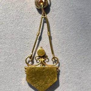 Puede incluir: Colgante de oro con dise&ntilde;o geom&eacute;trico. El colgante presenta un peque&ntilde;o lazo en la parte superior, conectado a un marco triangular con elementos decorativos. La parte inferior del colgante tiene forma de escudo.
