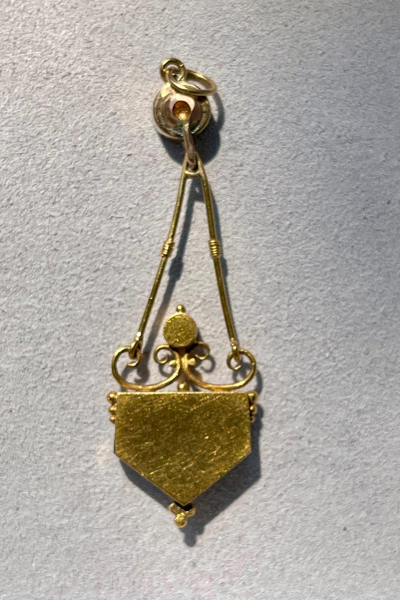 Puede incluir: Colgante de oro con dise&ntilde;o geom&eacute;trico. El colgante presenta un peque&ntilde;o lazo en la parte superior, conectado a un marco triangular con elementos decorativos. La parte inferior del colgante tiene forma de escudo.