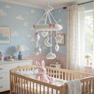 May include: A white baby mobile with pink bows and hanging shapes, including stars, clouds, and a crescent moon, hangs above a wooden crib. A pink stuffed bunny sits inside the crib. The nursery has a light blue wall with cloud patterns.