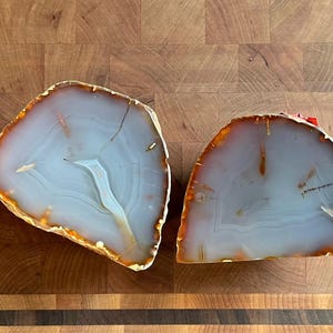 May include: Two polished agate slices with a light blue and white interior and a golden-orange edge. The natural stone has a banded pattern and is displayed on a wooden surface. These decorative pieces are ideal for home decor.