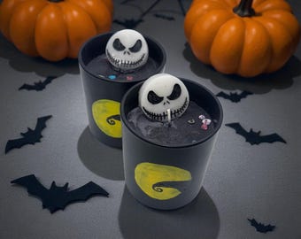 Könnte beinhalten: Zwei schwarze Halloween-Kerzen mit einem Jack-Skellington-Gesicht obenauf und einem gelben Monddesign. Zwei orangefarbene Kürbisse und schwarze Fledermaus-Ausschnitte sind ebenfalls im Bild. Die Kerzen stehen auf einer grauen Oberfläche.
