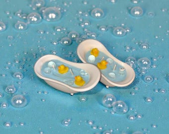 May include: Two miniature bathtubs filled with blue liquid and yellow rubber duckies. The bathtubs are white with a light blue interior. The background is a light blue surface with bubbles.