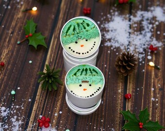 Pode incluir: Dois recipientes brancos com desenhos de bonecos de neve. Os rostos dos bonecos de neve têm cobertura verde para o chapéu, um nariz de cenoura e um sorriso pontilhado. O fundo apresenta azevinho, luzes e um pinha.