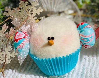 May include: A white felt snowman ornament with a blue cupcake liner, red and blue fabric accents, and black eyes. The snowman is wearing a blue and white patterned fabric hat.