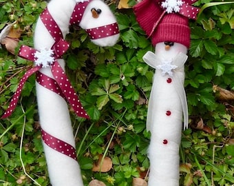 May include: Two handmade Christmas decorations, one shaped like a candy cane and the other like a snowman. The candy cane is white with red and white polka dot ribbon. The snowman is white with a red hat and white snowflake. Both decorations are made of felt.
