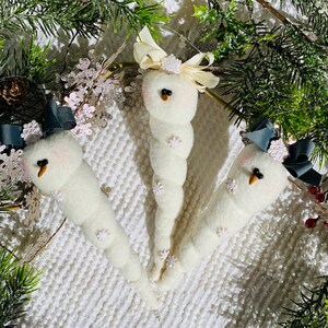 May include: Three white felt snowman decorations with black eyes and orange noses. Each snowman has a different coloured ribbon on top. The snowmen are on a white fluffy background.