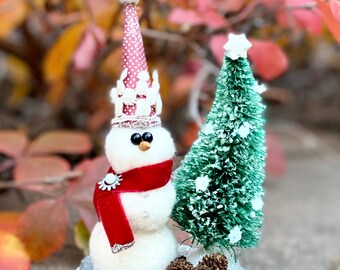 May include: A white felt snowman wearing a red and white hat and a red scarf, standing next to a small green artificial Christmas tree with white snow-like accents.