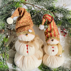 May include: Two handmade snowman ornaments with fabric hats and red bead accents. The snowman on the left has a brown and red patterned hat and a brown ribbon. The snowman on the right has a red and yellow striped hat and a brown ribbon.