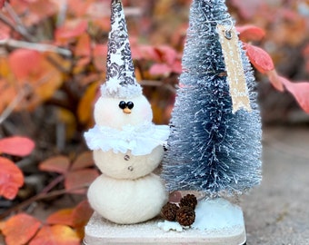 May include: A white snowman with a black and white patterned hat and a white ruffle collar stands next to a blue bottle brush Christmas tree with a brown tag that says "Let it Snow". The snowman and tree are on a white and black base.