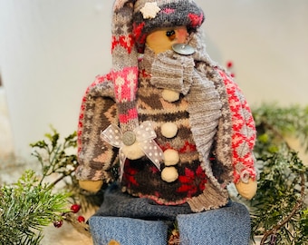 May include: A handmade snowman doll wearing a gray and pink knitted sweater, a gray scarf, and a pink and white knit hat with a white snowflake. The doll is sitting on a white surface with green pine branches and red berries.