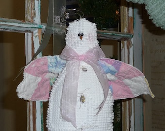 Può includere: Una statuetta di angelo bianca con un fiocco rosa e ali di stoffa patchwork. L'angelo ha un'aureola e un naso a bottone marrone.
