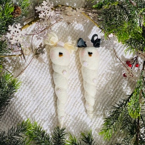 Puede incluir: Dos adornos de muñeco de nieve de fieltro blanco con ojos y boca negros. Un muñeco de nieve tiene un lazo blanco y el otro un lazo negro. Los muñecos de nieve están colgados sobre un fondo blanco esponjoso.