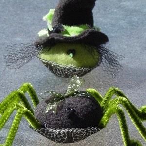May include: A green witch with a black hat and a black spider with green legs. Both are made of fabric and are sitting on a gray surface.