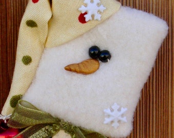 May include: A white felt snowman ornament with a yellow felt nose, black button eyes, and a white felt snowflake. The snowman is wearing a yellow fabric hat with red and green polka dots and a white felt snowflake.