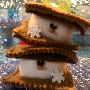 May include: Two stacked felt s'mores with white marshmallow centres, brown graham cracker sides, and white snowflake embellishments. The marshmallows have black button eyes and brown felt noses.