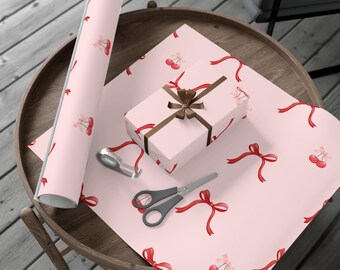 May include: A roll of light pink wrapping paper and a rectangular gift box wrapped in the same paper, both featuring a repeating pattern of red cherries with pink bows and red ribbons. A pair of silver scissors lies on the paper.
