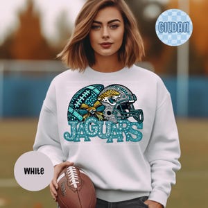 May include: A white long-sleeve sweatshirt featuring a graphic print of a football and a helmet with teal and gold accents. The design includes a leopard head logo and the text JAGUARS in a teal, glitter-style font. A brown football is held.