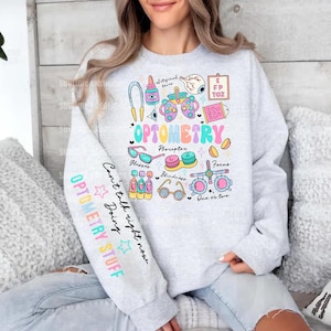 May include: A light gray crewneck sweatshirt featuring a colorful graphic print related to optometry. The design includes various eye care icons, the word OPTOMETRY, and text on the sleeve reading Can't talk right now doing OPTOMETRY STUFF.