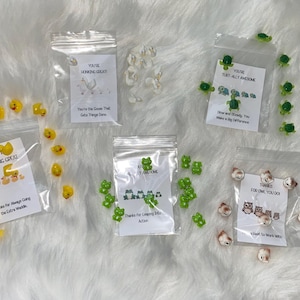 May include: A collection of small, clear plastic bags filled with various animal-shaped beads. The bags contain yellow duck, white goose, green frog, turtle, and owl beads. Each bag has a small card with a pun and a related image.