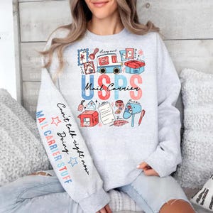 Puede incluir: Una sudadera gris claro de cuello redondo con un estampado gráfico de un camión de correo, paquetes y cartas con el texto USPS Mail Carrier. La manga izquierda muestra el texto Can't talk right now Doing MAIL CARRIER STUFF con estrellas.