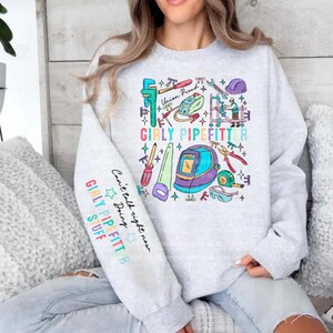 May include: A light gray crewneck sweatshirt featuring a colorful graphic print of various construction tools, a welding helmet, and the text GIRLY PIPEFITTER. The left sleeve includes the text GIRLY PIPEFITTER STUFF and Can't talk right now.