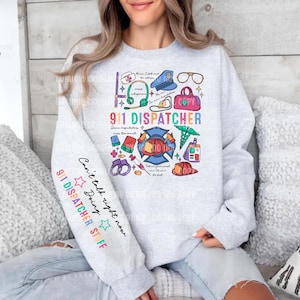 Puede incluir: Una sudadera gris claro de cuello redondo con un estampado gráfico colorido en el frente y la manga. El diseño frontal incluye el texto 911 DISPATCHER rodeado de iconos de auriculares, gorra de policía, símbolos médicos y esposas. La manga dice 911 DISPATCHER STUFF.