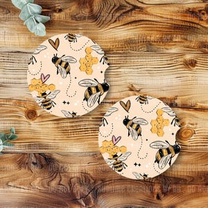 May include: Two round car coasters with a bee-themed design. The coasters feature a light beige background with illustrations of bees, honeycombs, and hearts. The bees are black and yellow, and the hearts are pink and brown.
