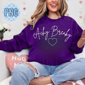 Puede incluir: Una sudadera morada con la frase "Achy Breaky" en escritura blanca y un corazón. La sudadera se combina con vaqueros azules. Se ve una almohada rosa en forma de corazón con la palabra "HUG".