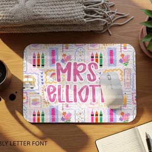 May include: A rectangular mouse mat with a white background and a colourful pattern of crayons, flowers, and school-themed illustrations. The name "MRS ELLIOTT" is written in large, pink, bubbly letters. A computer mouse rests on the mat.