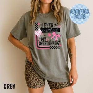 Puede incluir: Camiseta gris Comfort Color con el texto "I even overthink my overthinking" en un diseño rosa y negro. El diseño incluye un corazón, estrellas y un lazo a cuadros.